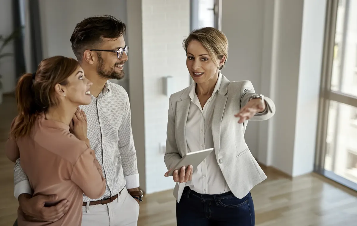 Una pareja junto a una mujer en una vivienda mientras ésta señala con la mano